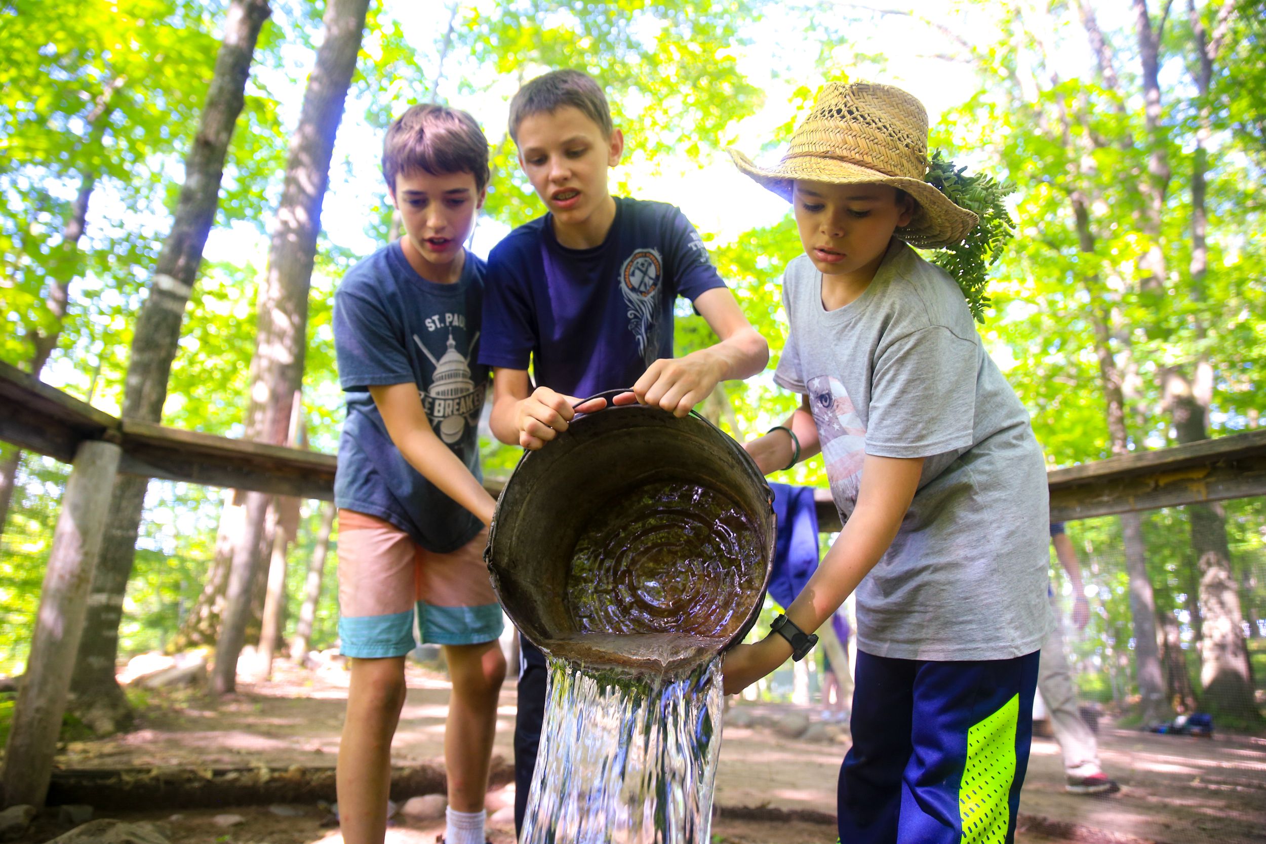 Group activity involving water being poured from a large pan in a wooded camp setting.