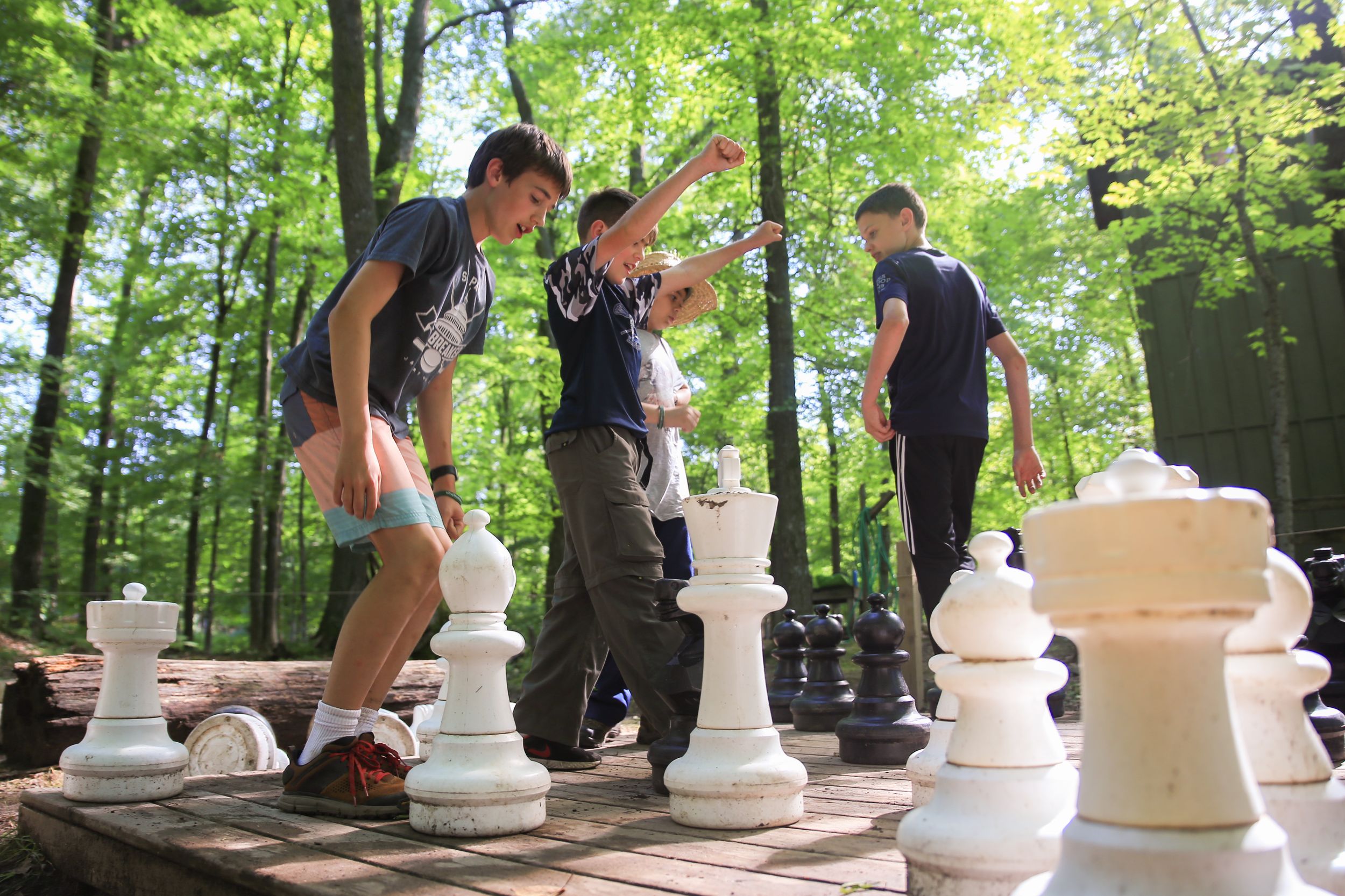 Kids playing with giant chess pieces on an outdoor board surrounded by trees.