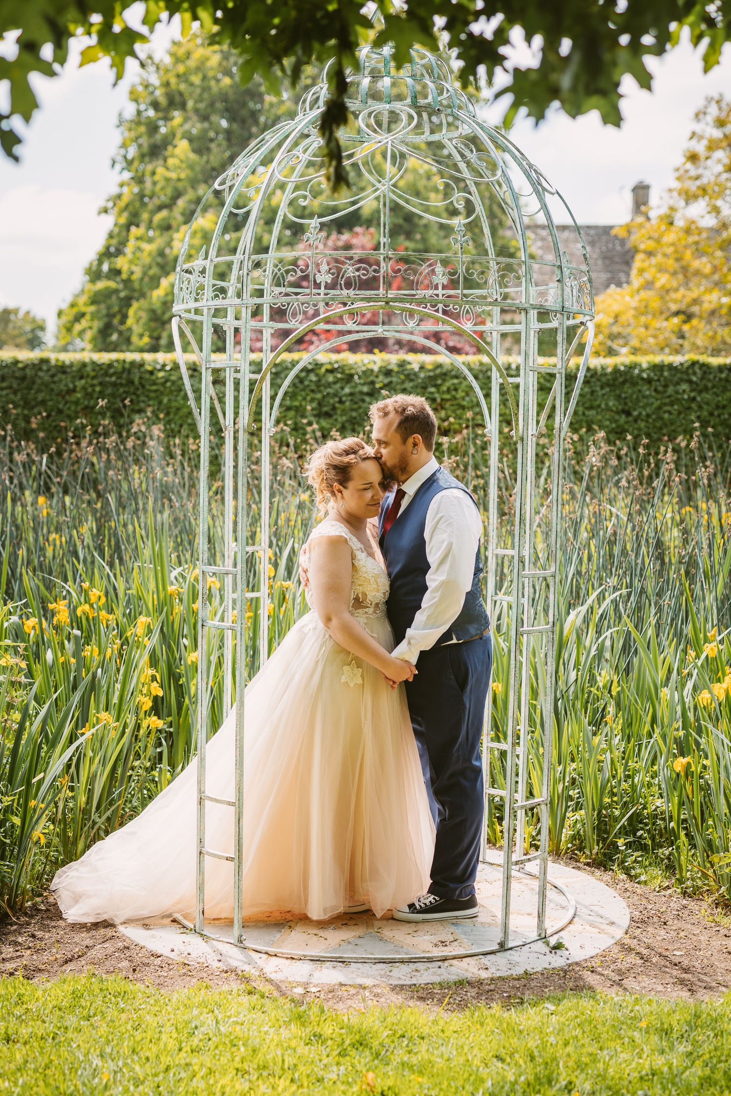 Wedding couple embraces inside ornate white garden gazebo surrounded by yellow iris flowers.