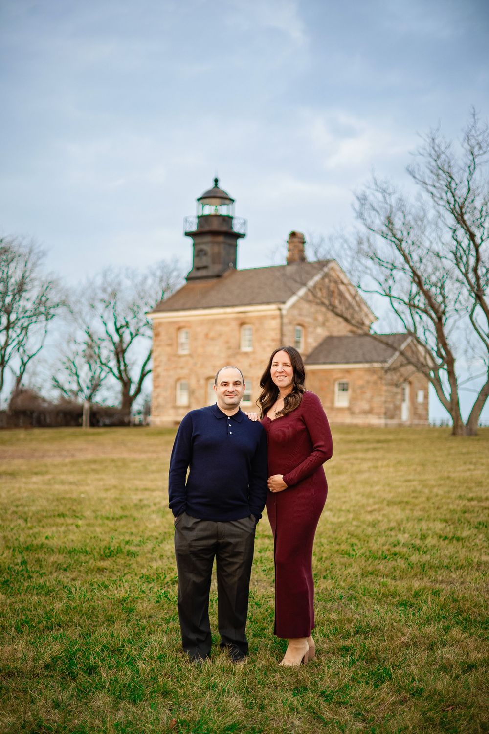 A couple poses together on a grassy hillside with a historic lighthouse and brick building in the background.
