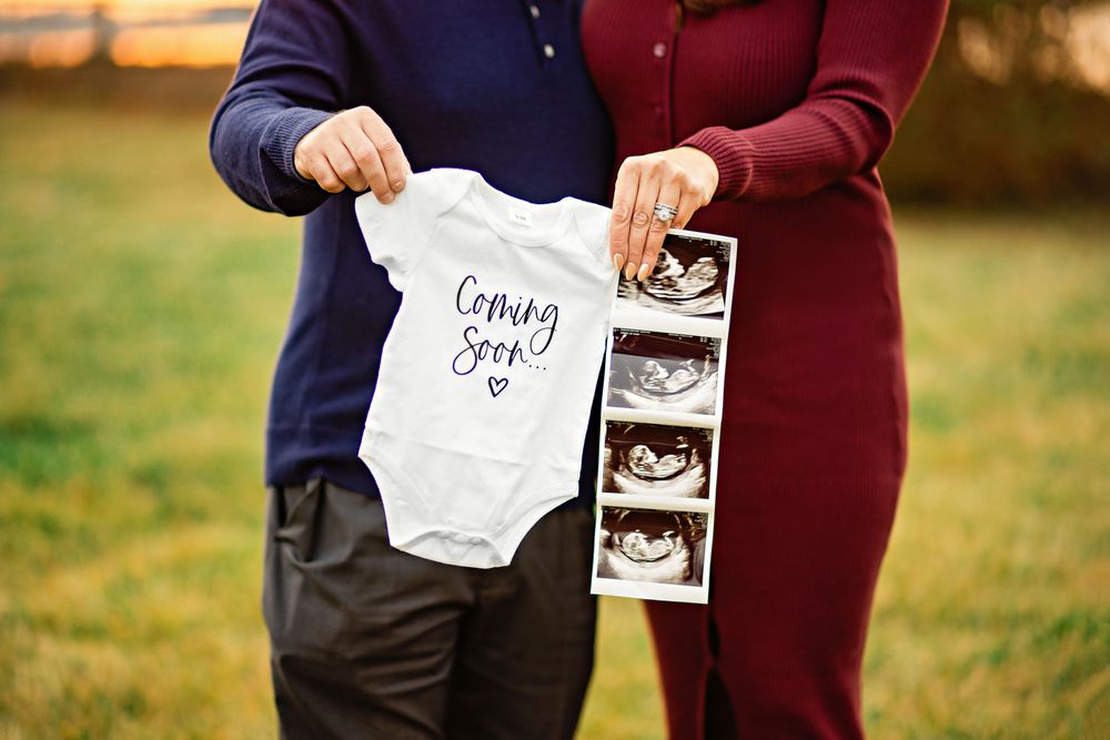A couple holding ultrasound photos and a sign during a pregnancy announcement photo session at sunset.