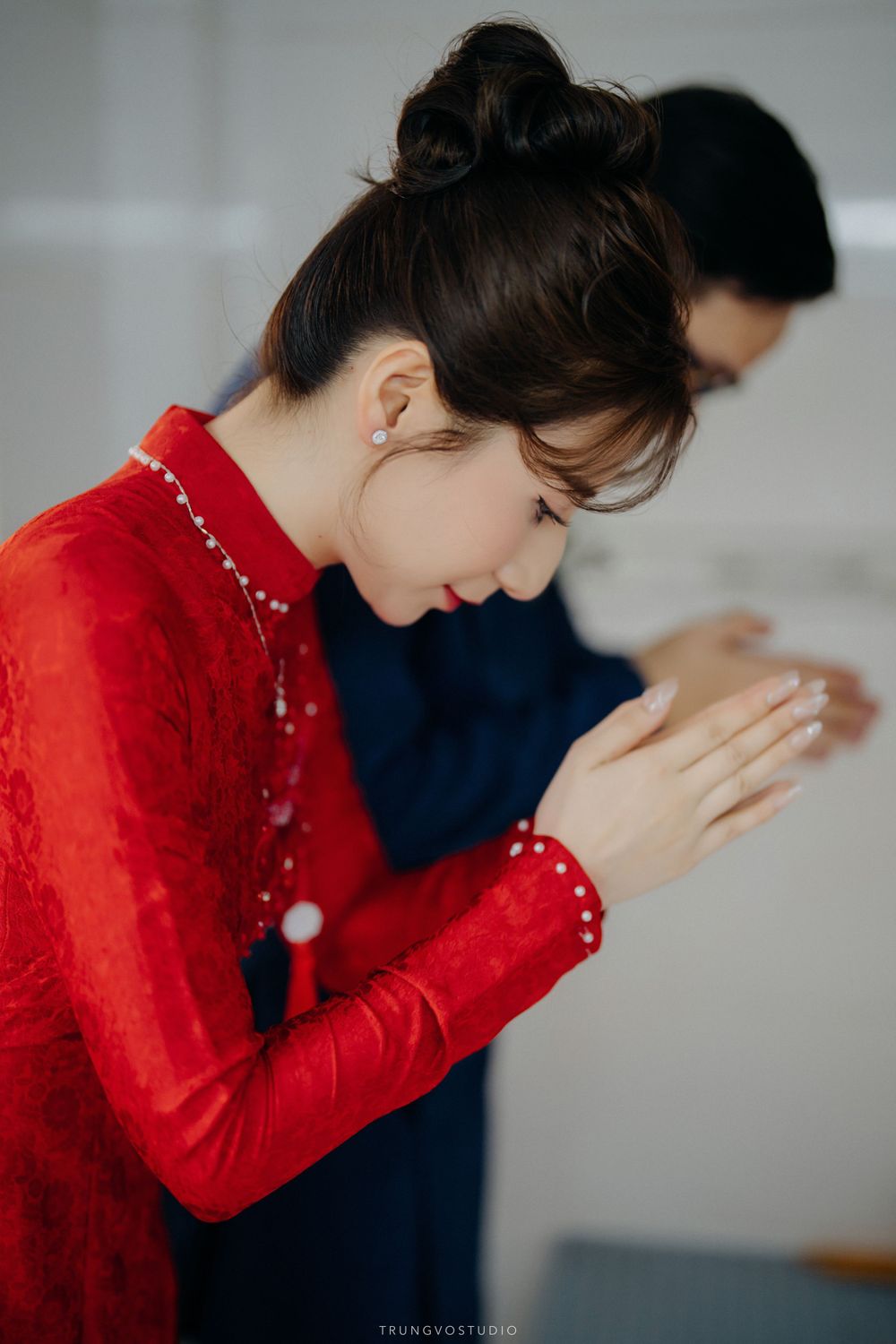 Young woman in red ao dai bows respectfully with hands clasped during Vietnamese wedding ritual.
