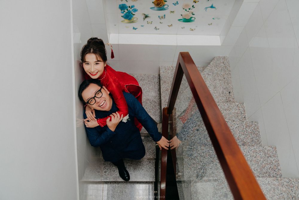 Smiling couple in traditional Vietnamese wedding attire pose together on marble staircase.