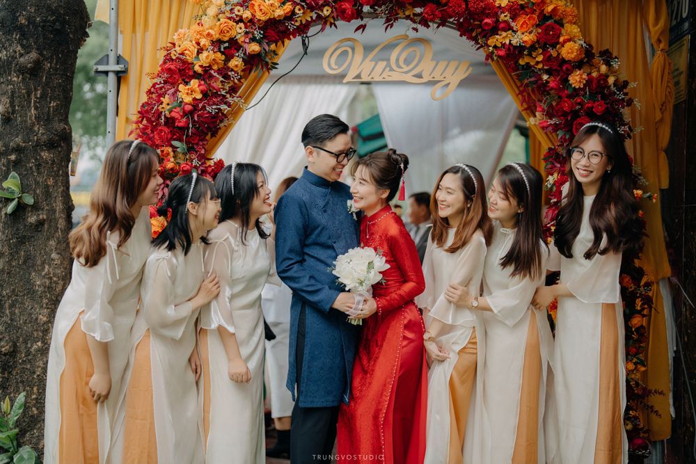 Bride and groom in Vietnamese ao dai share tender moment surrounded by bridesmaids at floral arch.