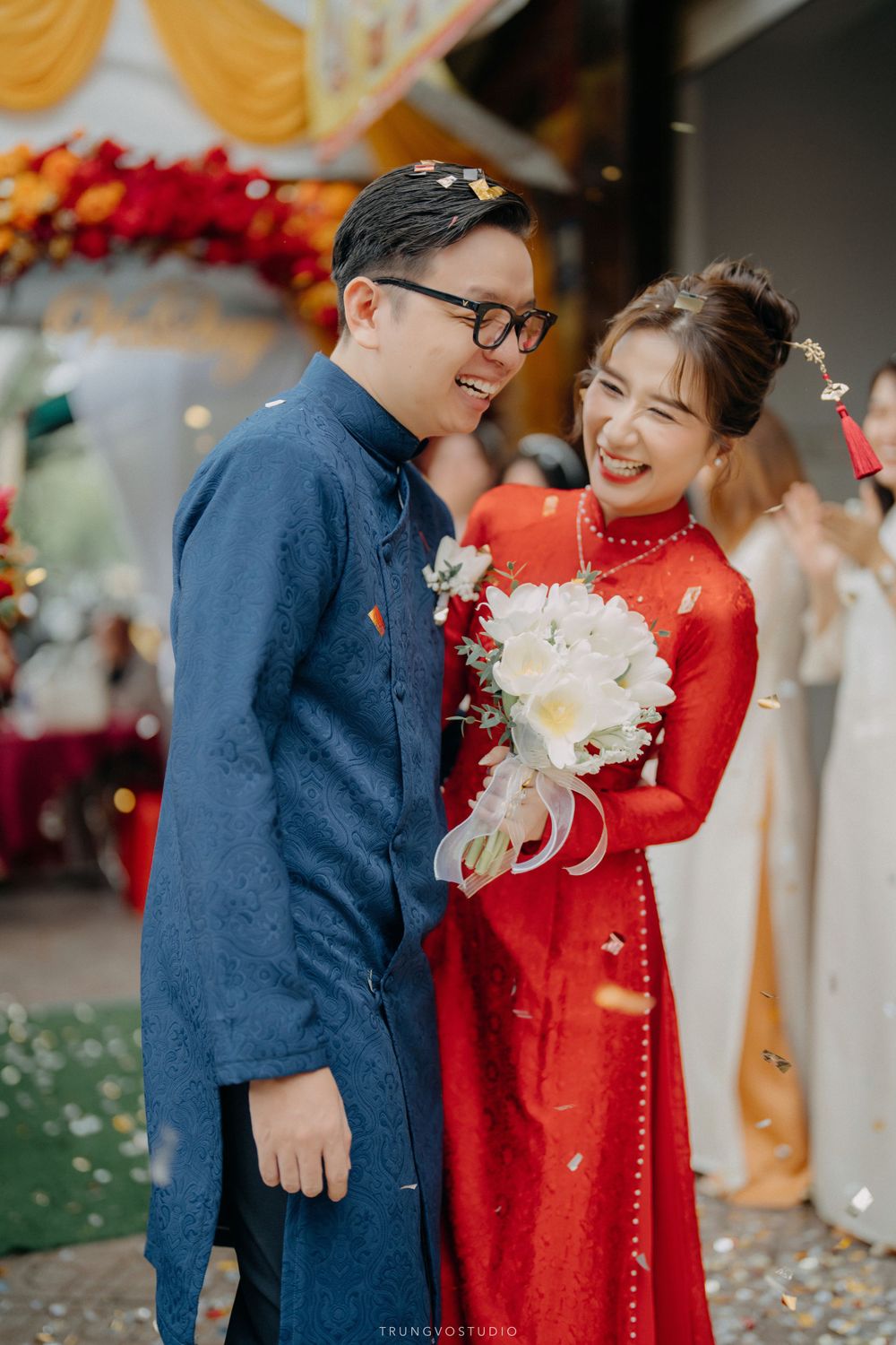 Joyful Vietnamese couple in ao dai laugh together as confetti falls during outdoor wedding.