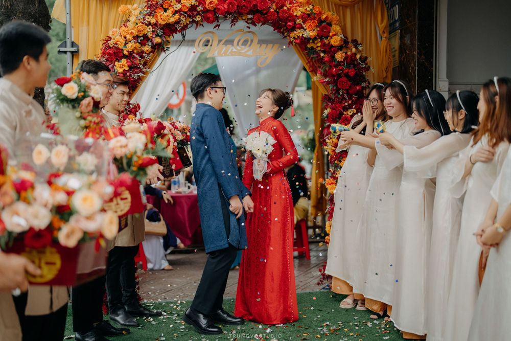 Vietnamese bride in red ao dai and groom in blue walk through floral arch at traditional wedding ceremony.