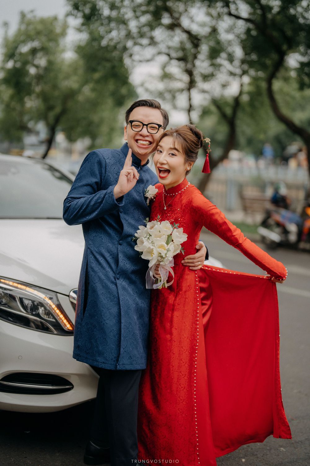 Vietnamese couple in traditional ao dai wedding attire pose joyfully beside a white Mercedes car.