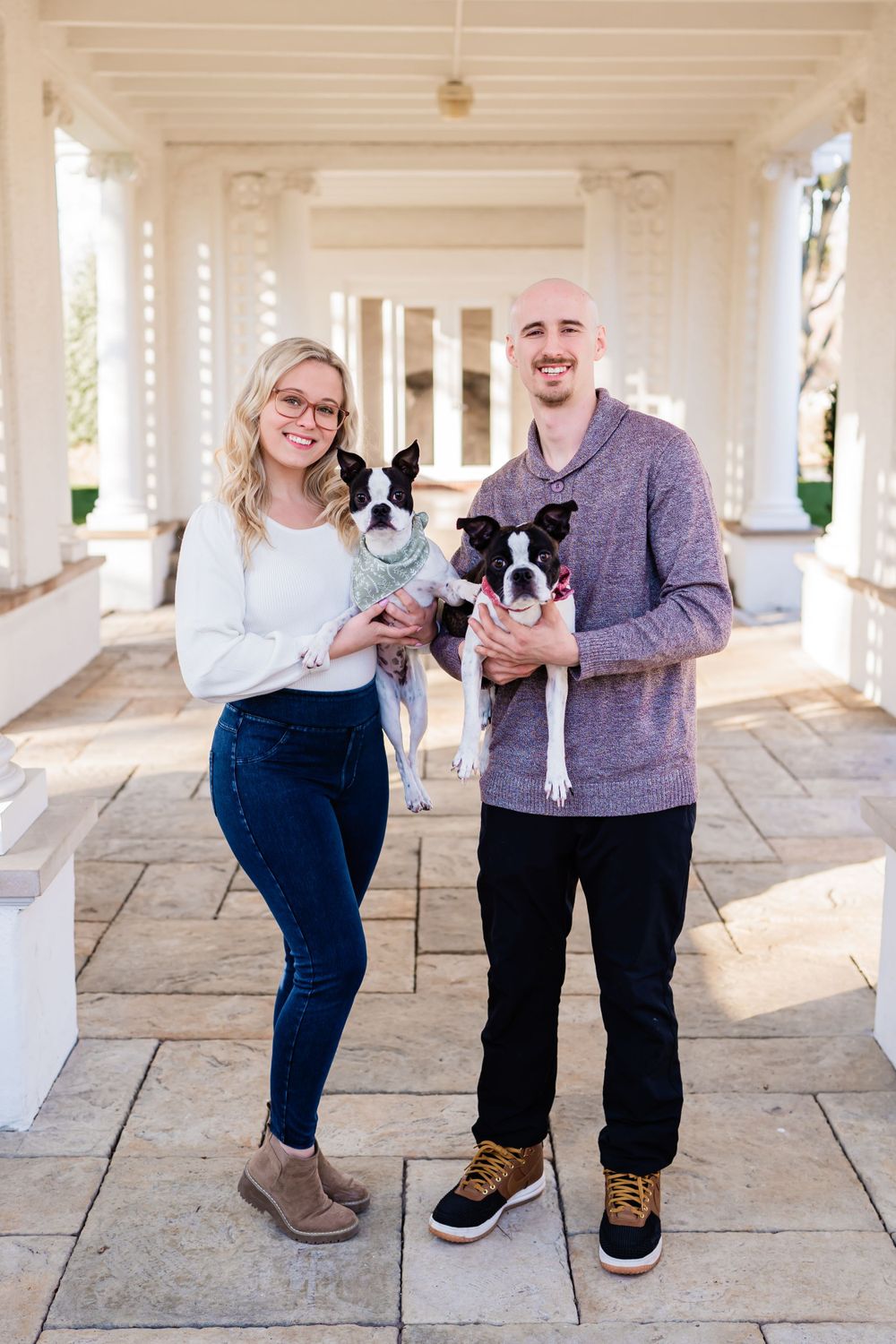A couple stands on a columned porch holding their two Boston Terrier dogs while posing for a casual photo.