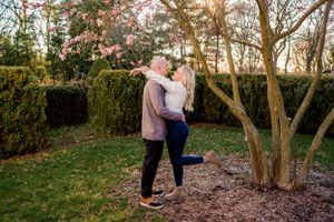 A romantic couple embraces in a garden setting with tall hedges and trees at sunset.
