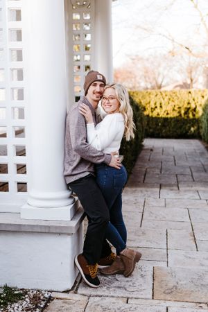 A romantic couple embraces while standing against a white pillared gazebo on a stone patio during autumn.