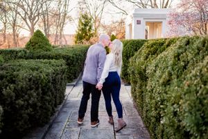 A tender moment shared between a couple in a garden walkway lined with manicured hedges at sunset.