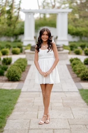 Young girl in white mini dress and sandals poses on stone path in elegant garden with manicured hedges and white pergola.