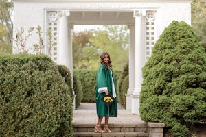 Graduate in green cap and gown stands before white garden pergola surrounded by manicured hedges holding yellow flowers.