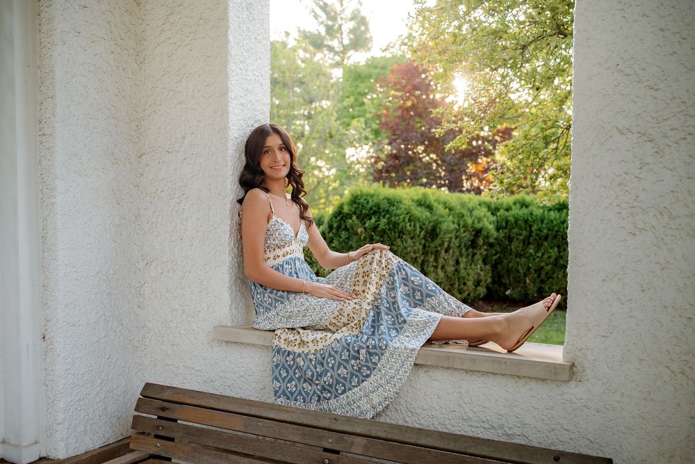 Young woman in floral maxi dress sitting gracefully on a window ledge with sunlit greenery in the background.