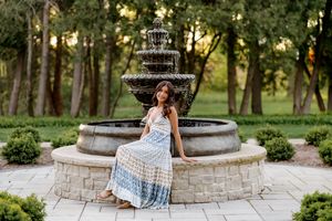 Young woman in a blue boho maxi dress poses by an ornate stone fountain in a lush green garden.