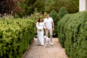 A couple in white outfits walking through a garden path with tall green hedges during a photo session.