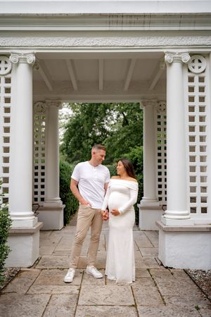 A couple stands under an ornate white garden pergola with Ionic columns and lattice details during a maternity photoshoot.