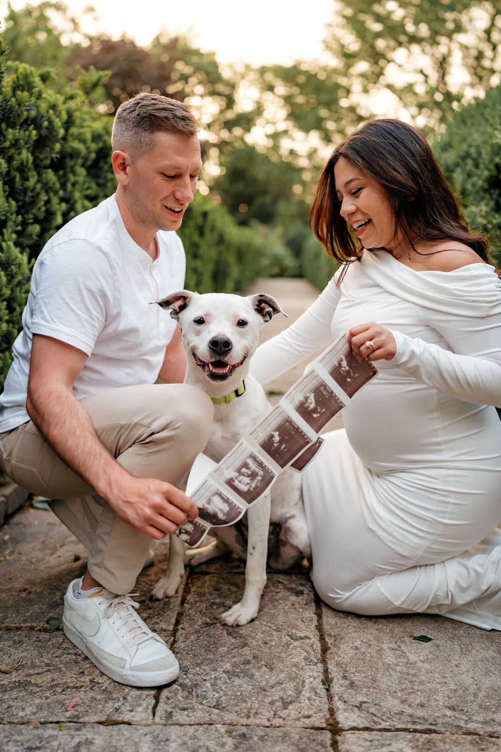 A couple sits with their white dog outdoors while looking at ultrasound photos during a pregnancy announcement photoshoot.