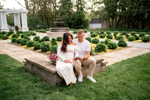 A couple sits together on a stone wall in a manicured garden with boxwood hedges and white pergola.
