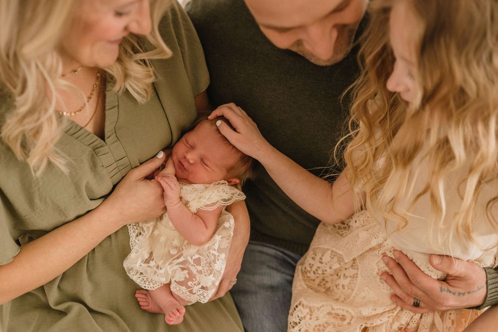 A newborn baby in lace clothing is lovingly cradled by adults in a warm, intimate indoor setting.