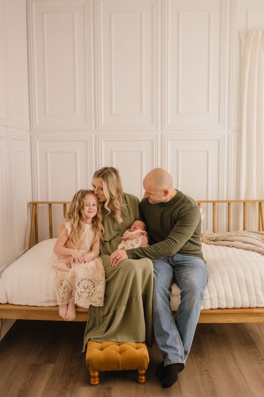 A cozy family portrait on a wooden bed frame with white paneled walls and warm lighting.