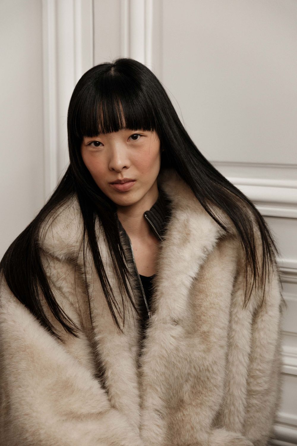 Fashion model wearing luxurious beige fur coat poses against white wall.