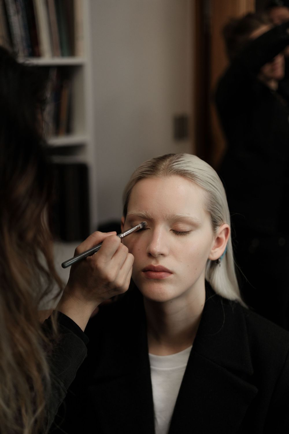 Makeup artist applying eyeshadow to model during beauty session with bookshelves in background.