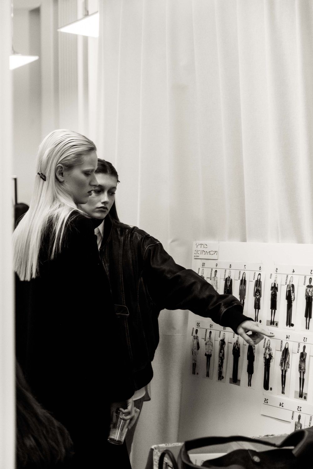 Two people reviewing fashion design sketches posted on wall in black and white.