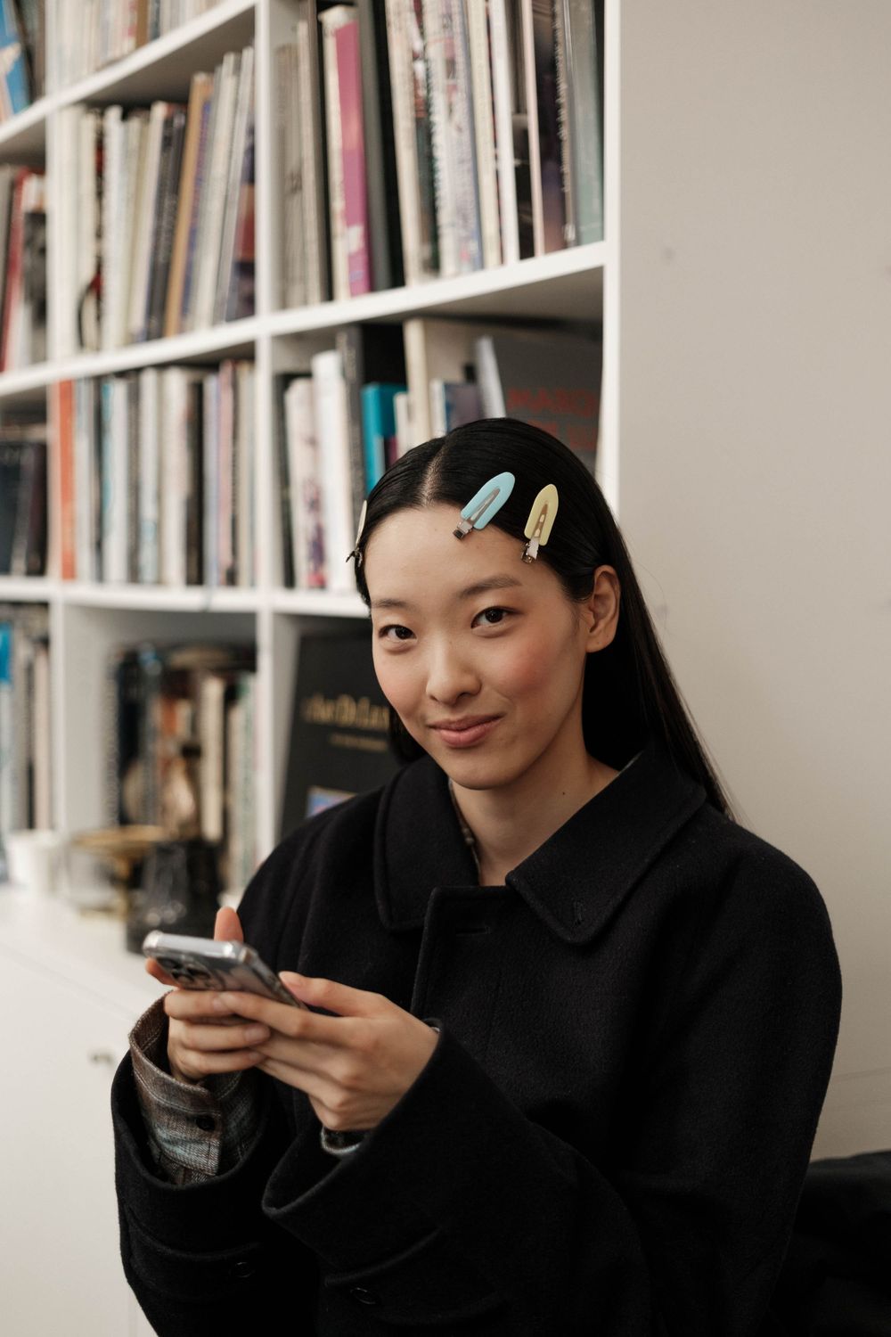 Young woman in black coat using smartphone while sitting near bookshelf.