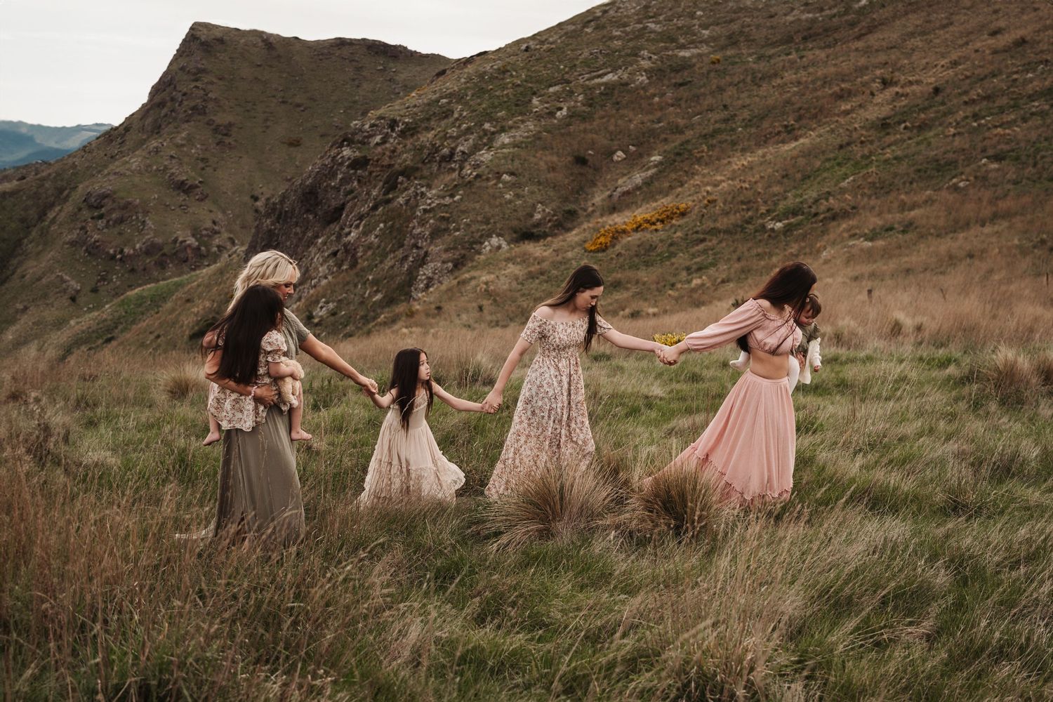 People in flowing dresses hold hands while dancing through tall grass on a hillside at sunset.