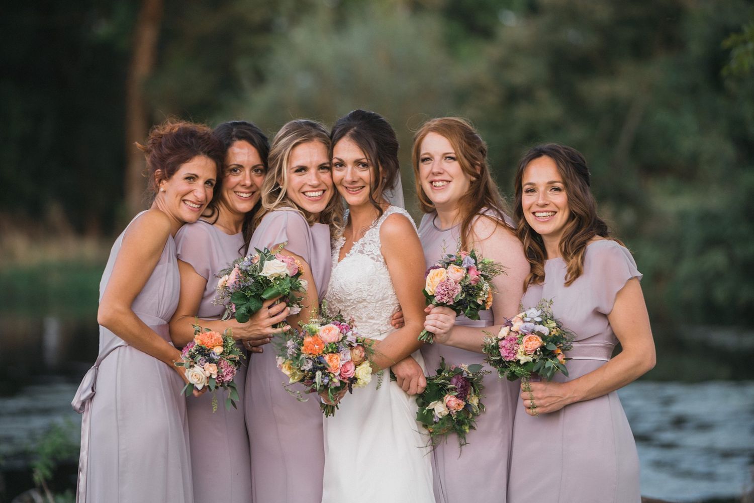 A bridal party poses together outdoors at The Farm At South Cove wearing lavender dresses and holding colorful bouquets by a river.