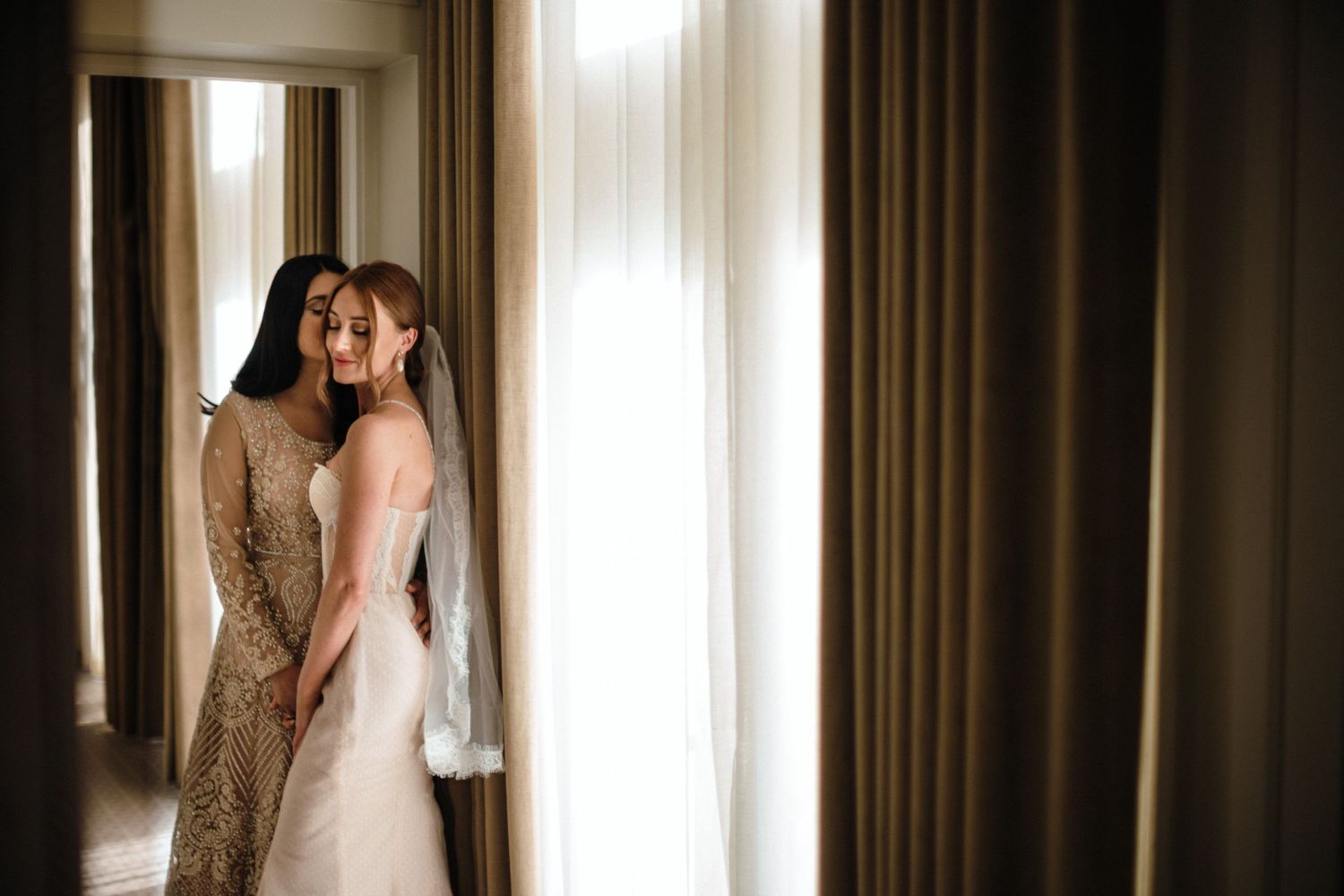 A bride in a white wedding dress stands with bride in an Indian wedding dress near a sunlit window with curtains at The Lord Nelson hotel.