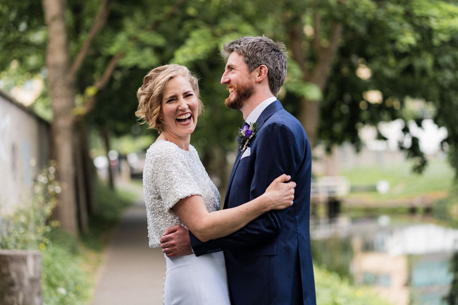 A couple embraces outdoors on their wedding day, wearing a lace dress and navy suit among trees in a park setting.