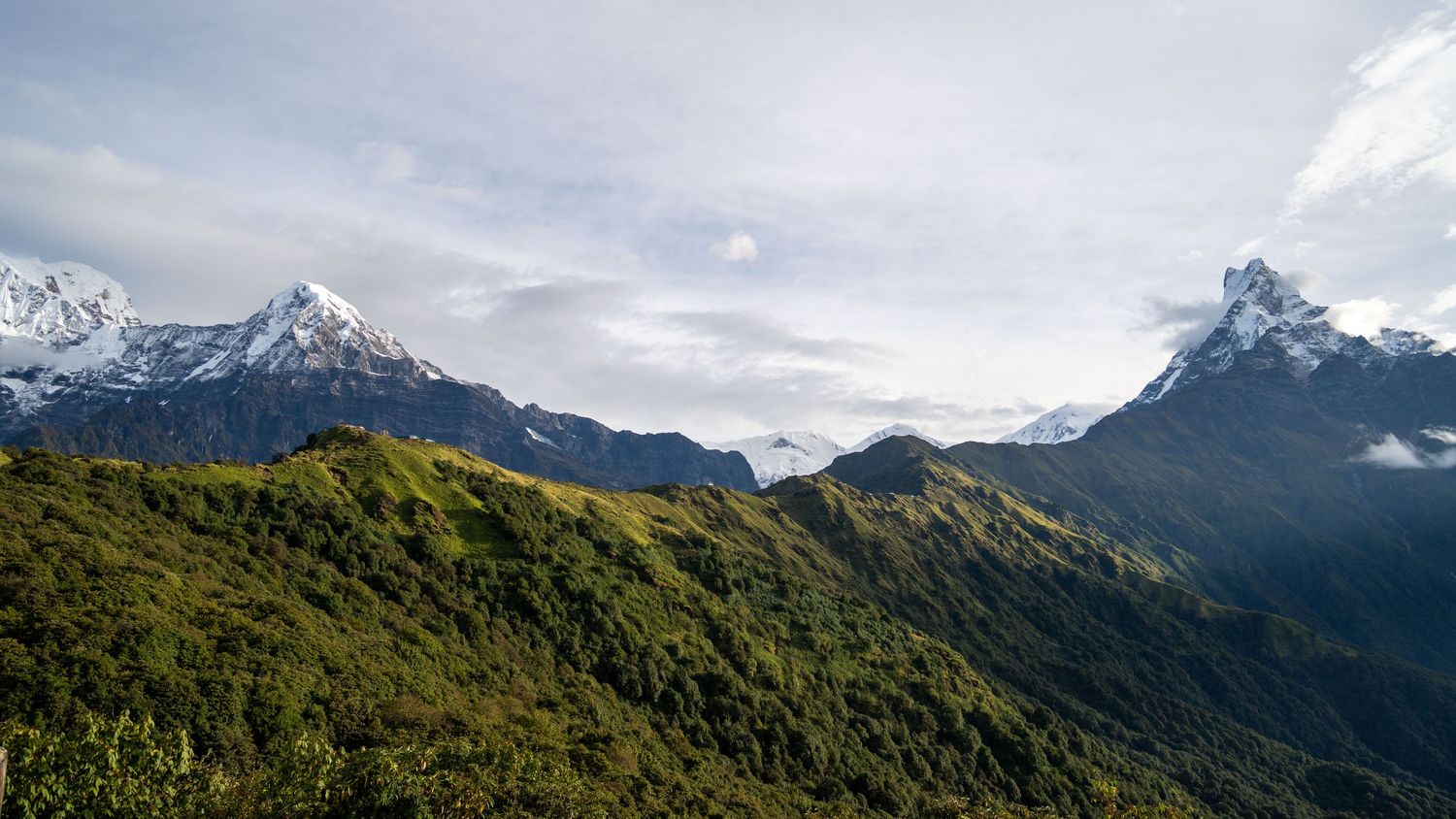 Panoramic mountain landscape with snow-capped peaks and green valleys under cloudy skies.