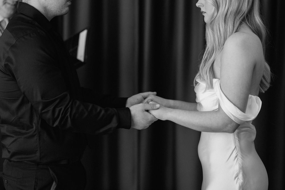 Black and white photo of a romantic couple holding hands during a wedding ceremony, the bride wearing an elegant white gown.