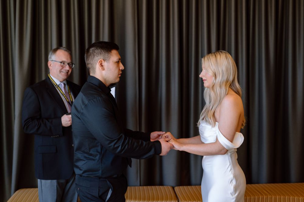 A couple holds hands during an indoor wedding ceremony with dark curtains in the background while an officiant observes.