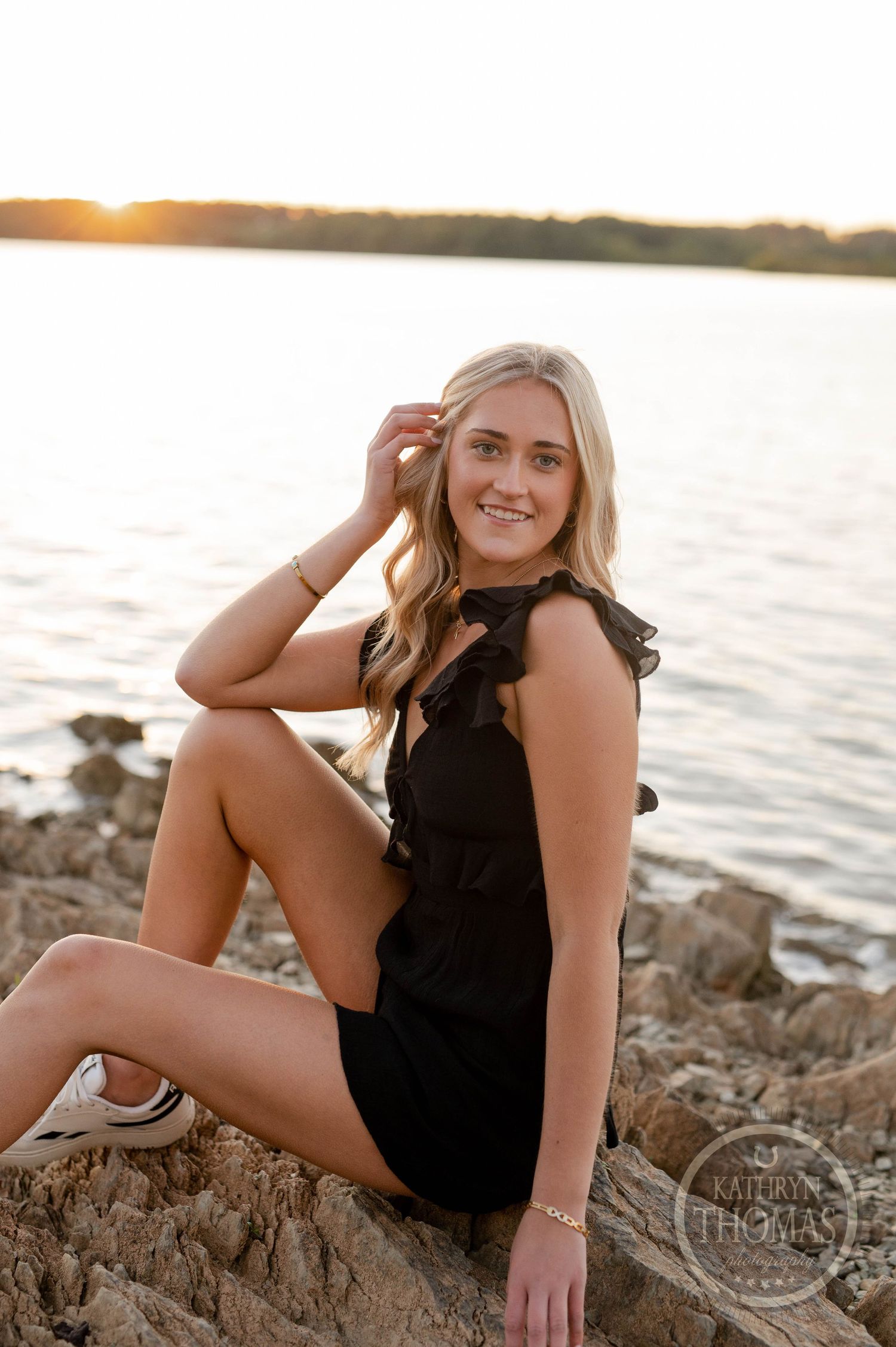 Summer portrait of someone in black dress sitting on rocky shoreline at sunset.