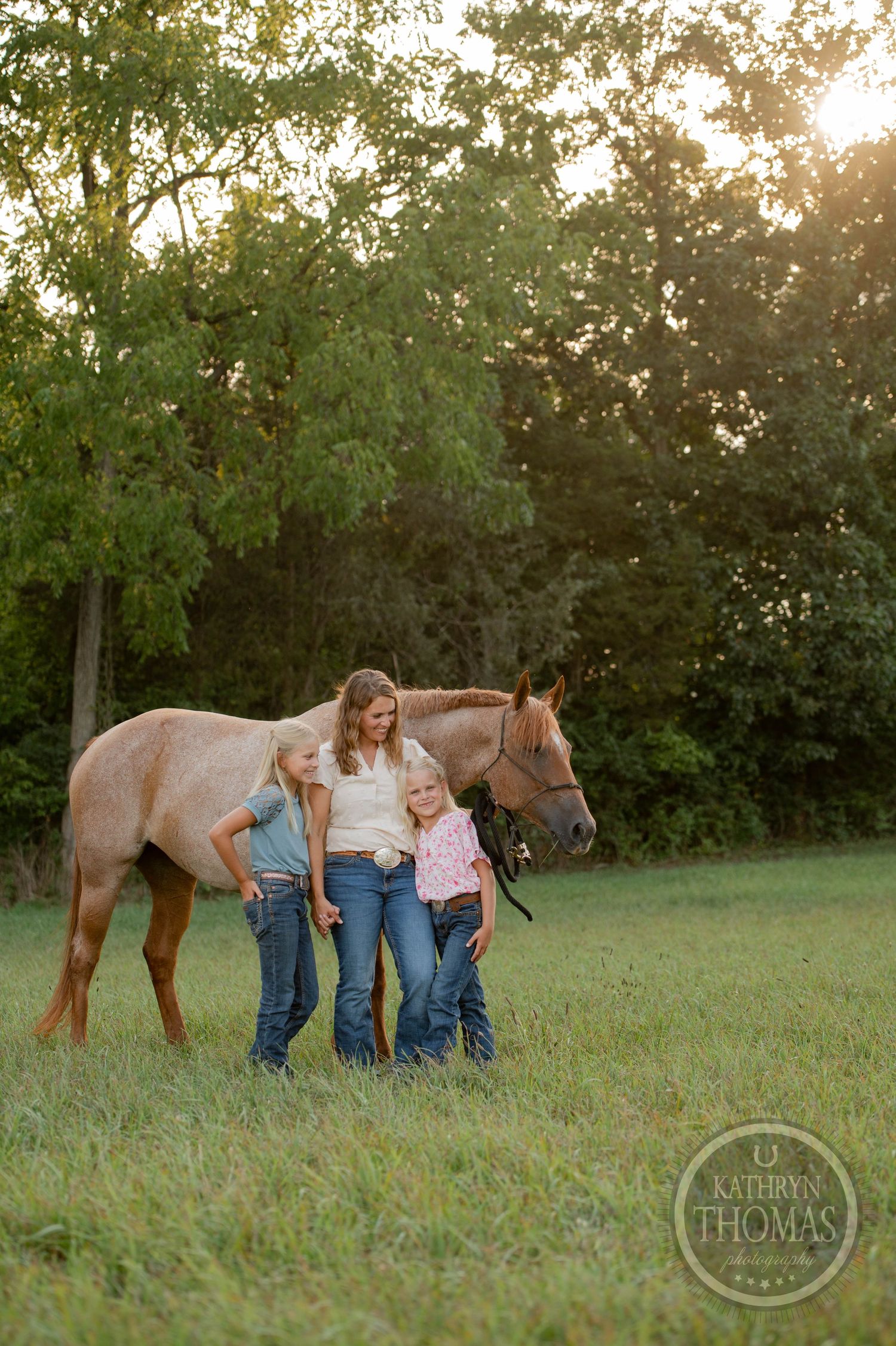Three people stand beside a brown horse in a serene pastoral setting during sunset.