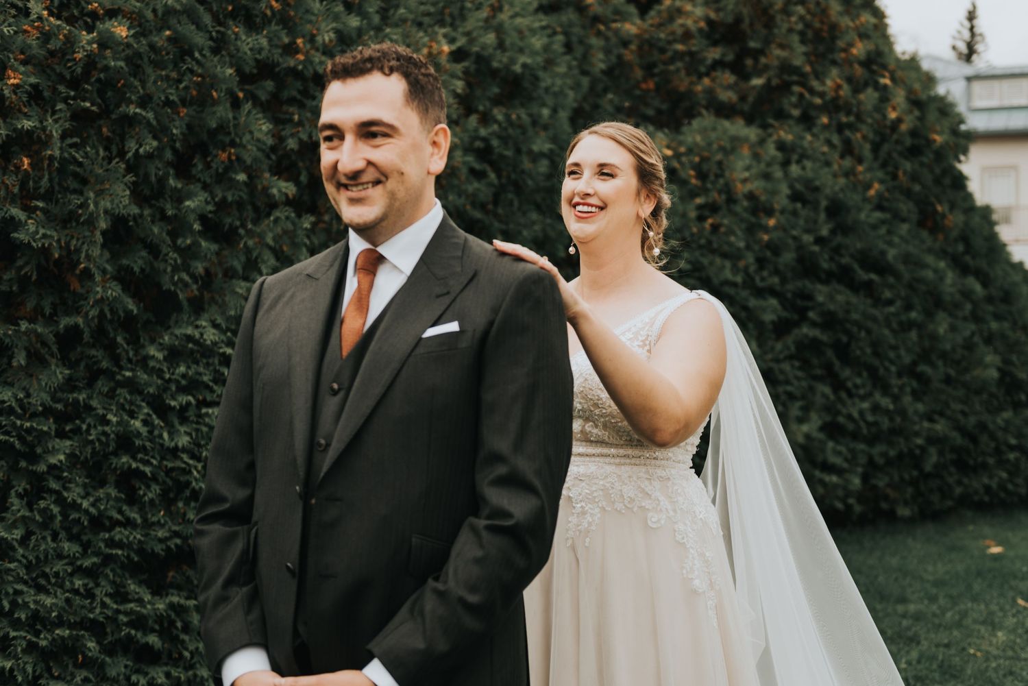 A bride in a white gown and veil smiles while standing behind her groom in a dark suit during their wedding first look.