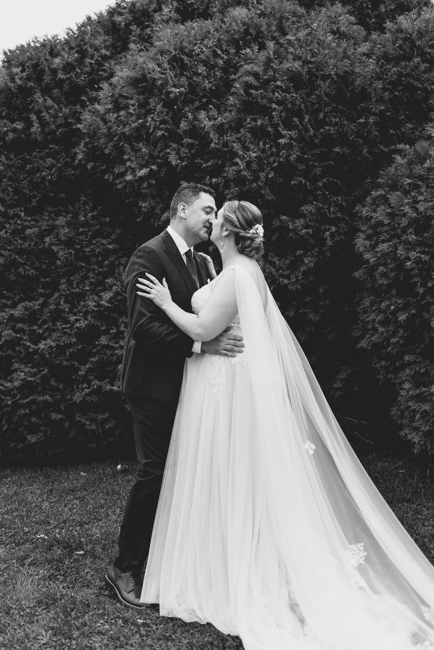 A black and white wedding photo shows a couple embracing outdoors in front of a lush garden hedge.