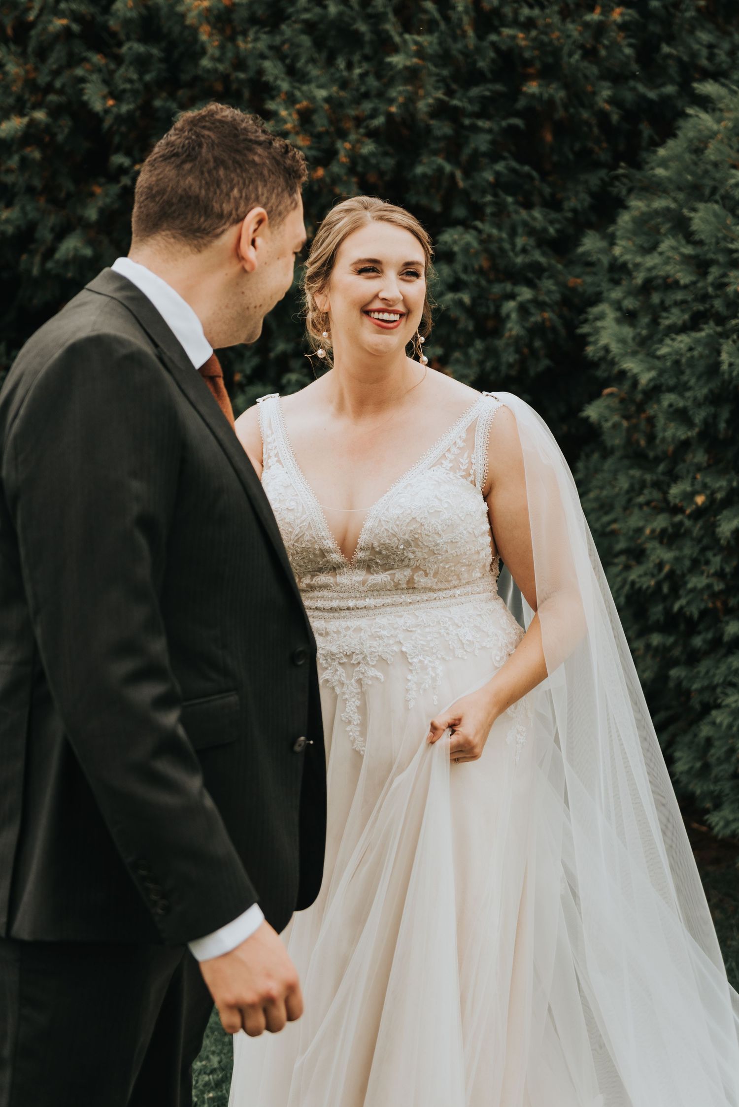 A romantic wedding moment captured with the bride in a flowing white gown and veil standing with her groom against greenery.