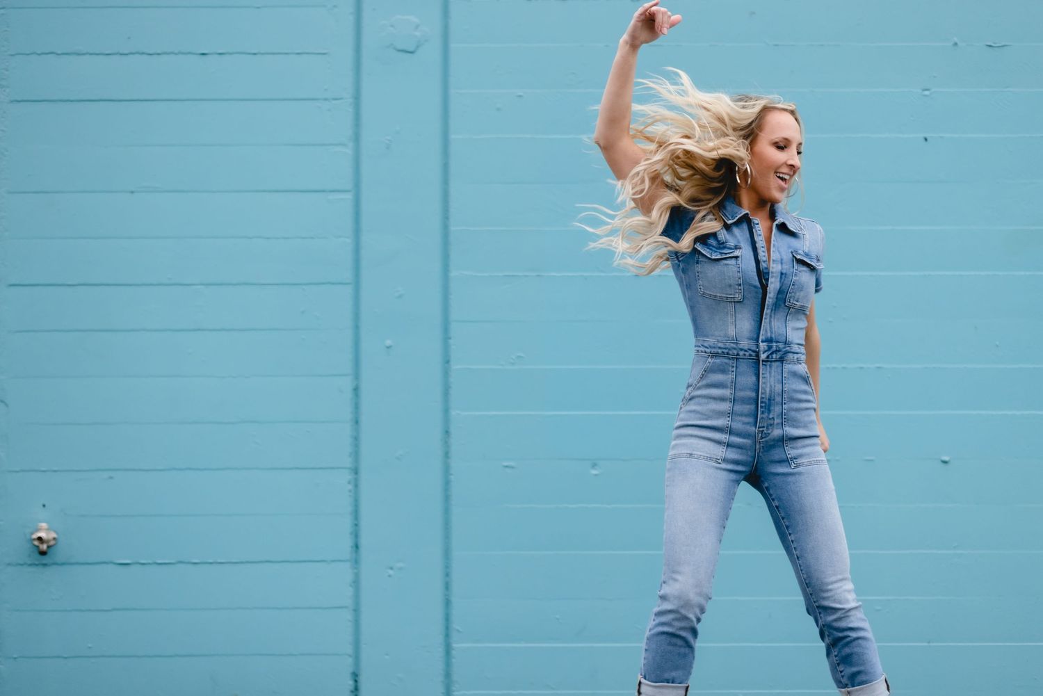 Energetic person in denim jumpsuit dancing and jumping in front of turquoise wall with flowing blonde hair.