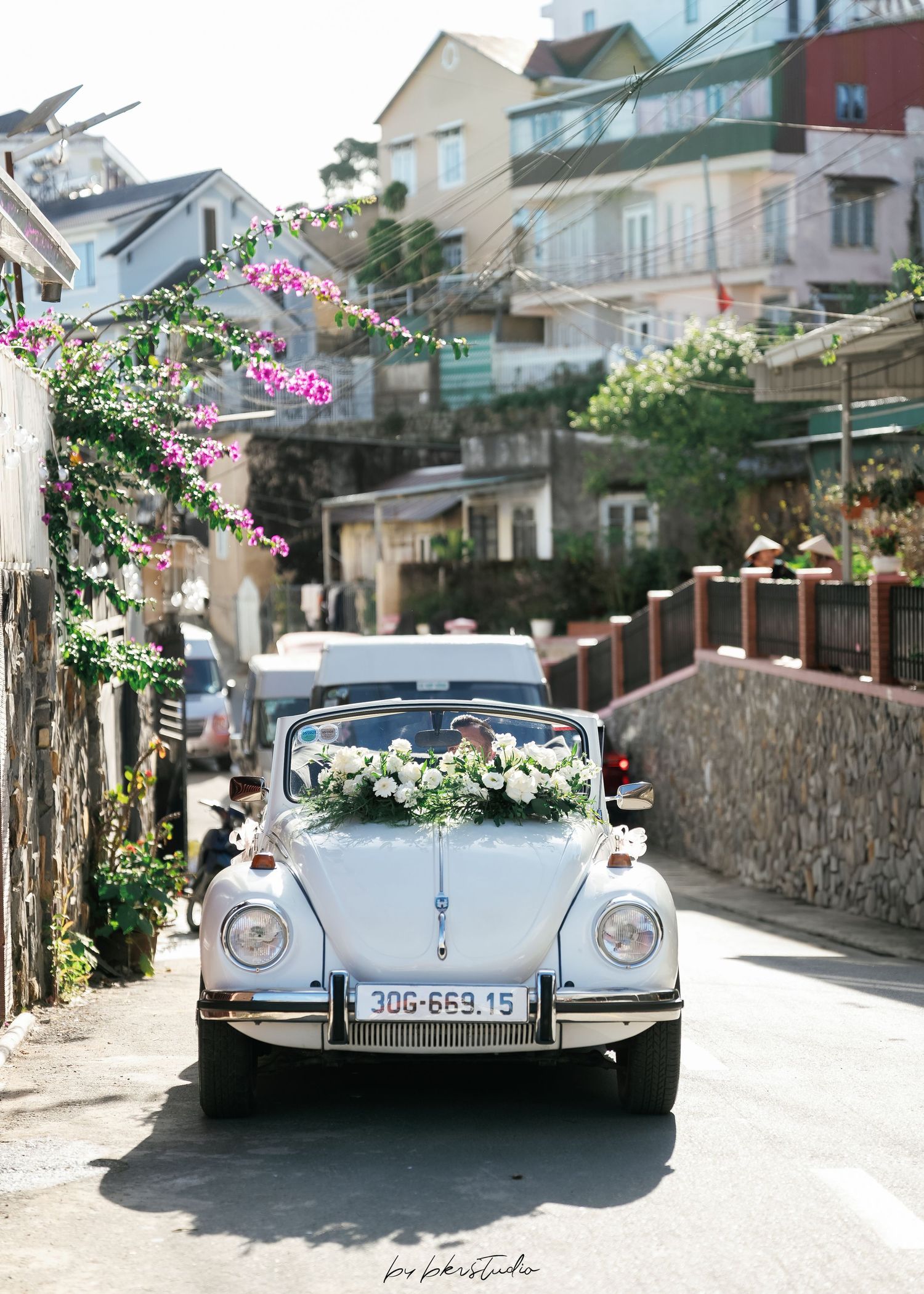 A vintage white Volkswagen Beetle decorated with flowers sits on a charming European street with hillside buildings.