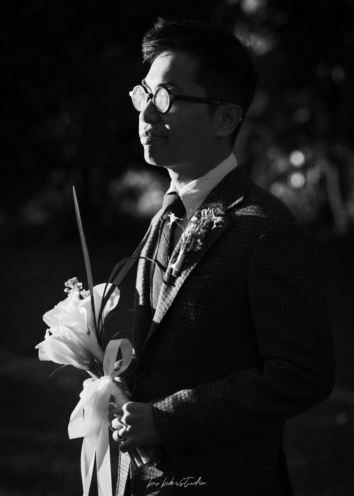 Black and white portrait of a person in a formal suit and glasses with a corsage in dramatic lighting.