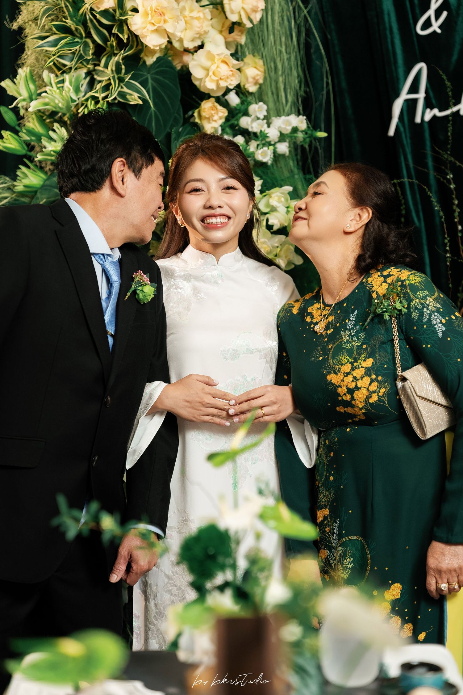 Three wedding guests share a joyful moment surrounded by floral decorations and a dark green backdrop.