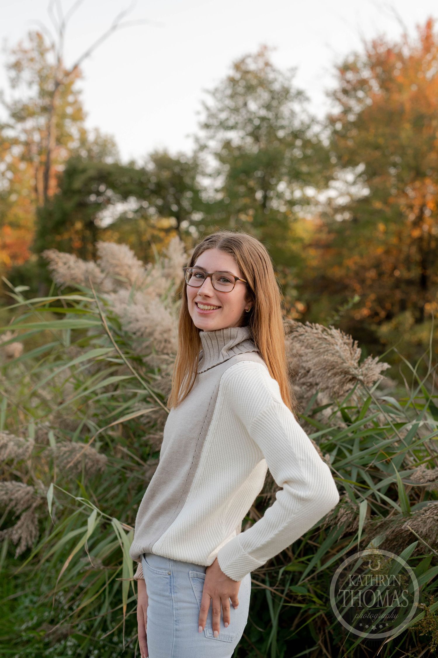 Portrait against autumn foliage showing someone in glasses and a cream turtleneck sweater.