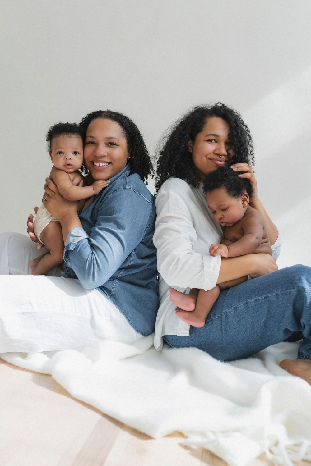 Two adults in casual denim sitting back-to-back on white bedding while holding infants.