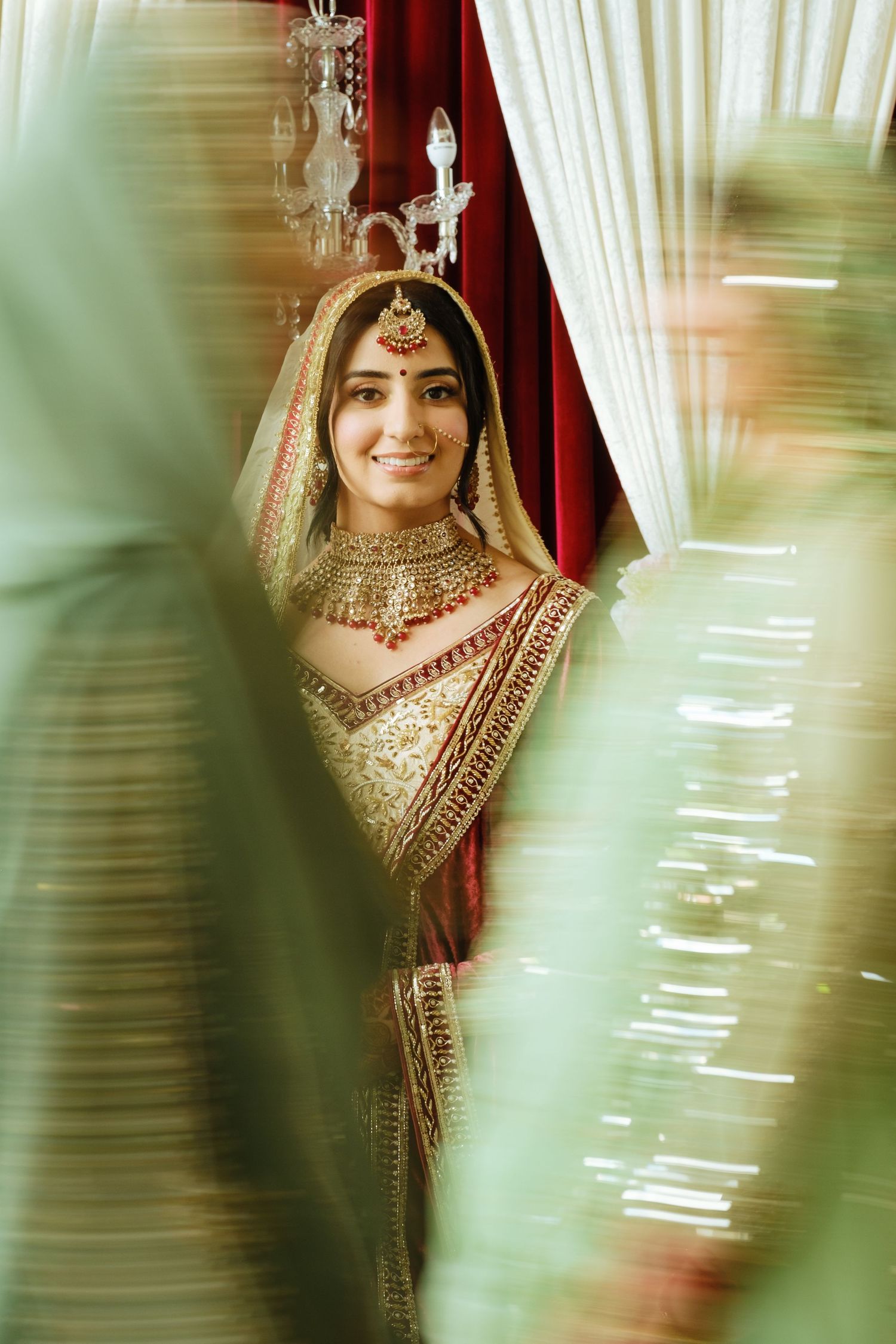Beautiful traditional Indian bridal attire with intricate gold jewelry and embroidered red lehenga captured through sheer curtains.