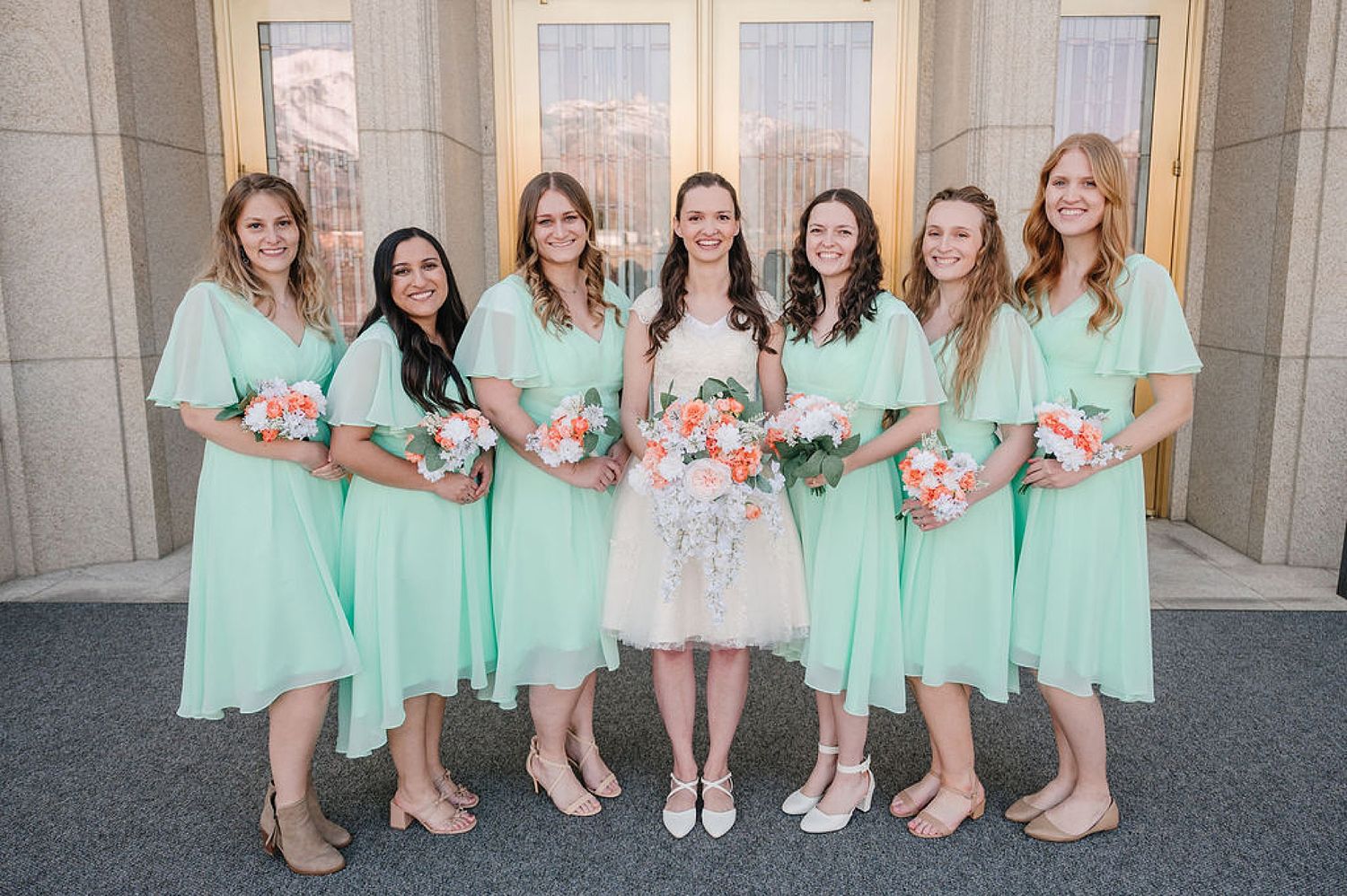 A bridal party wearing mint green dresses poses with pink bouquets alongside a bride in white at a church entrance.