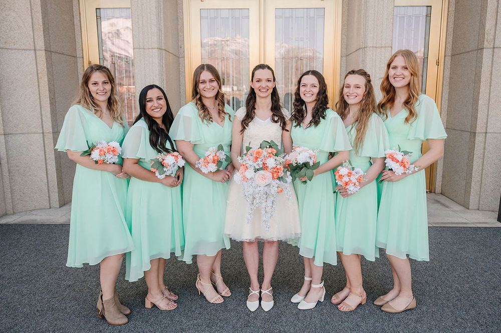 A bridal party wearing mint green dresses poses with pink bouquets alongside a bride in white at a church entrance.